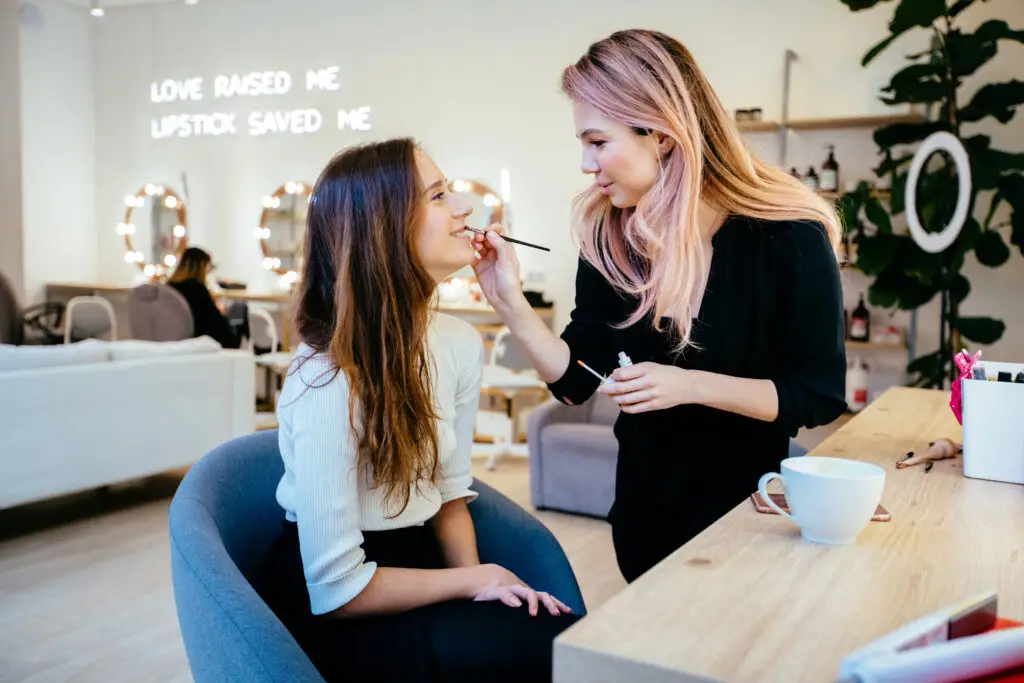 Friendly positive make up artist applying gloss on woman lips in new, expensive modern beauty salon interior. Shoot with real people model