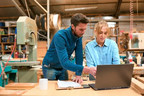 Male And Female Carpenters Working In Woodwork Workshop Using Laptop Together